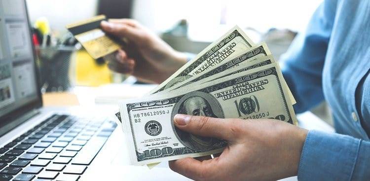 A person holding a stack of US dollar bills in one hand and a credit card in the other, sitting in front of a laptop computer. The scene suggests online shopping or financial transactions. The background includes a blurred-out office setting with office supplies.