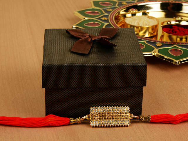 A black gift box with a brown ribbon is placed on a wooden surface. In front of the box, there is a decorative rakhi with red threads and an intricate gold centerpiece. A decorative plate with items for a Raksha Bandhan celebration is seen in the background.