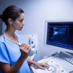 Woman in blue scrubs using an ultrasound machine, looking at the screen displaying an image.
