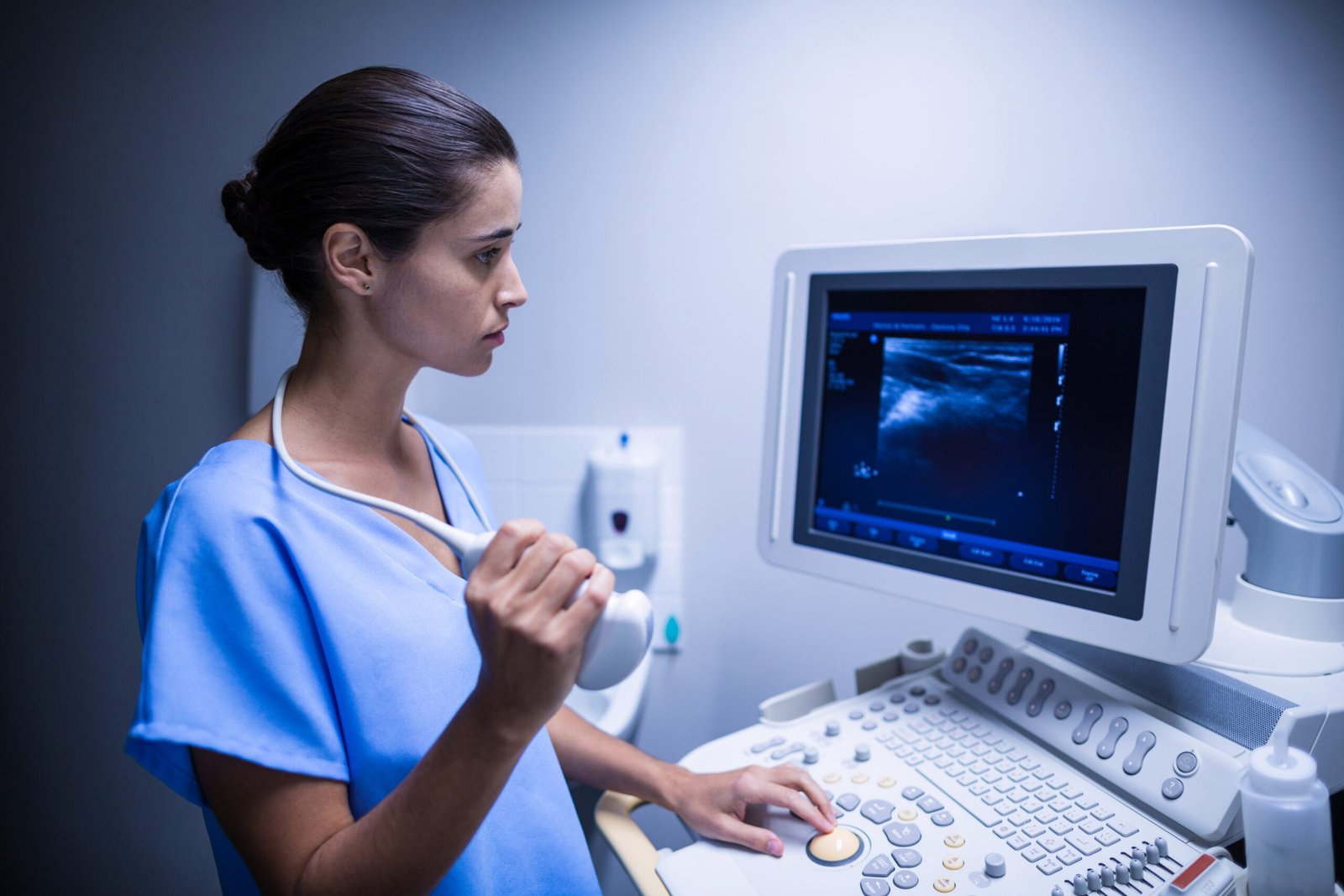 Woman in blue scrubs using an ultrasound machine, looking at the screen displaying an image.