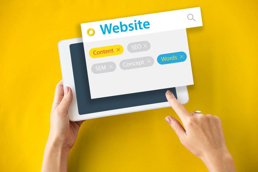 Hands hold a tablet with a floating search box overlay. The search box displays the word "Website" with keywords "Content" in yellow, "SEO" and "SEM" grayed out, "Concept" in gray, and "Words" in blue. The background is solid yellow.