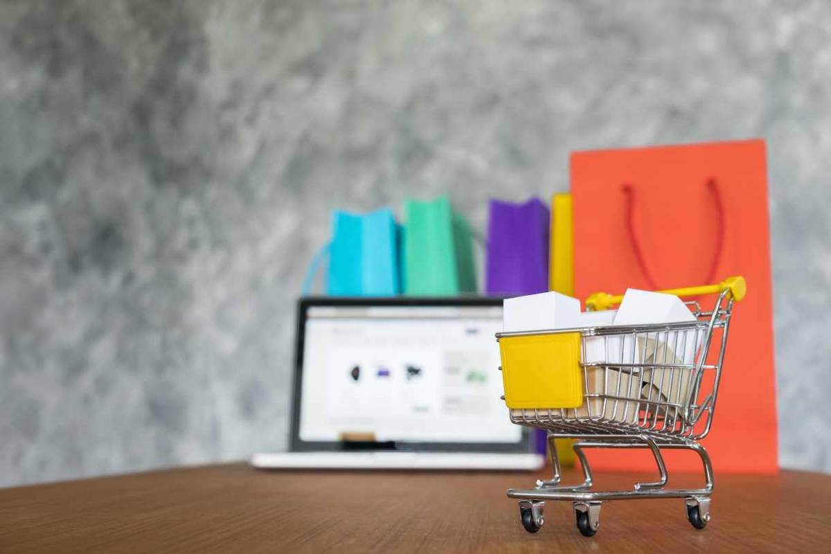 A miniature shopping cart filled with small boxes sits on a wooden surface. In the background, a laptop displaying a shopping website and several colorful shopping bags are visible. The scene depicts online shopping and e-commerce.