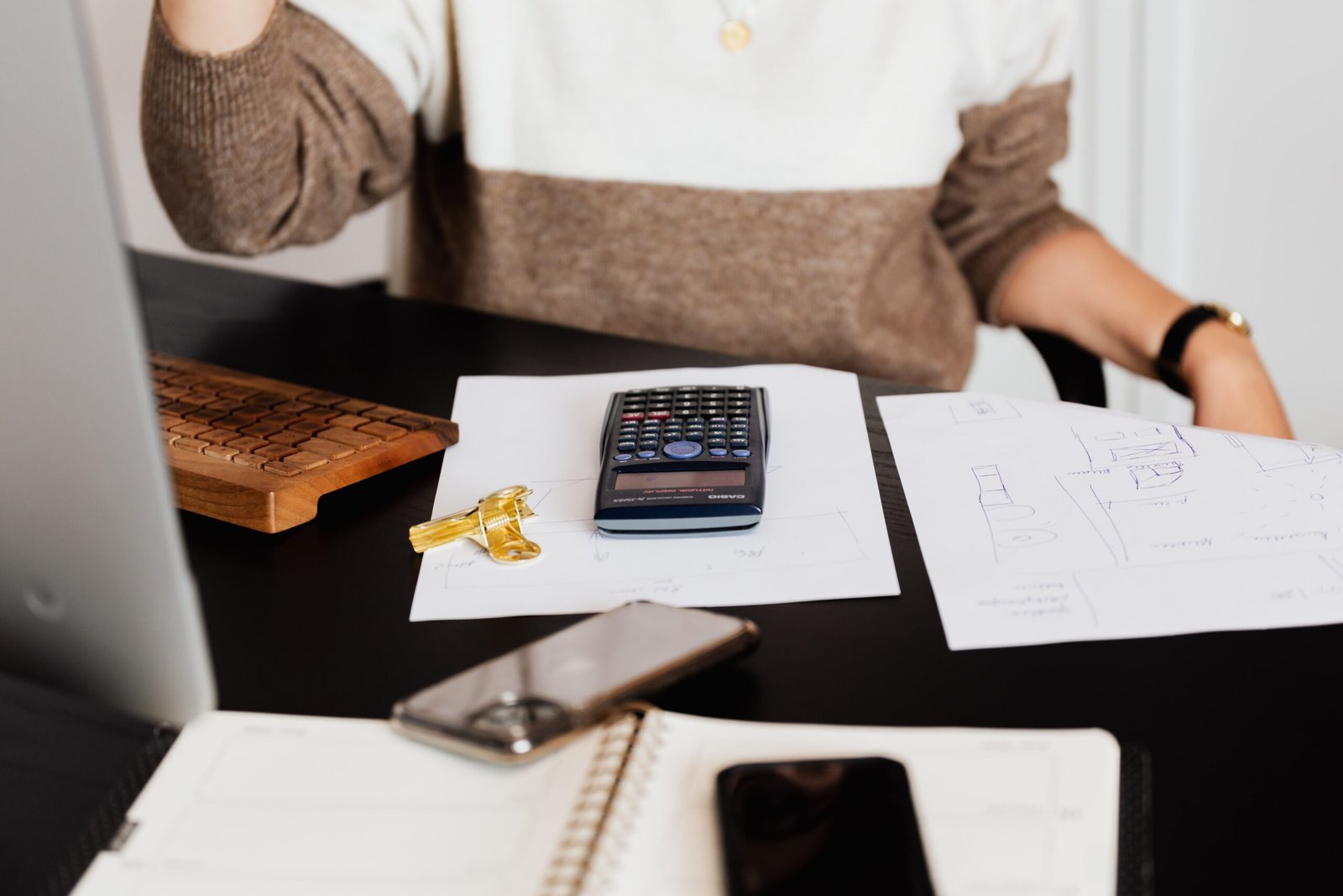 A person sitting at a desk with a brown and white sweater, partially visible. On the desk are a calculator, a wooden keyboard, keys, printed documents, and a smartphone. An open notebook and another smartphone are in the foreground.