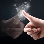 A hand pressing a glowing fingerprint icon on a transparent digital shield, symbolizing biometric security. The background is dark, with digital circuits and binary code, emphasizing the connection between technology and secure identity verification.