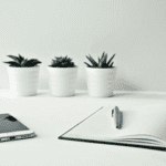 A white desk with a smartphone, an open notebook with a pen placed on top, and three small potted succulents arranged in a row against a light-colored wall. The arrangement appears neat and minimalist.