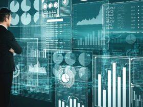A man in a suit stands with arms crossed, facing a large transparent screen filled with various data visualizations including charts, graphs, and statistics, against a dark, high-tech background with city lights visible in the distance.