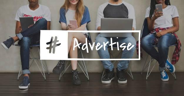 Four young adults sit in a row, using various devices like smartphones and a laptop. An overlay text reads "#Advertise," implying a focus on digital media or marketing. They are casually dressed and seated against a plain wall with a wooden floor.