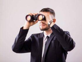 A man with short hair and a beard is wearing a dark suit and tie. He is holding a pair of binoculars up to his eyes and looking through them. The background is a plain, light gray color.