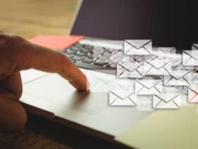 A person's hand is shown pointing towards a laptop keyboard. Several digital envelope icons, symbolizing emails, are overlaid on top of the image, giving the impression of multiple emails being sent. The background includes part of the laptop and workspace.