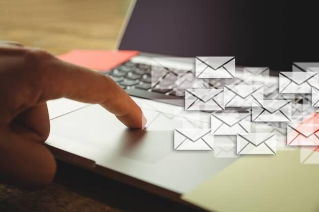 A person's hand is shown pointing towards a laptop keyboard. Several digital envelope icons, symbolizing emails, are overlaid on top of the image, giving the impression of multiple emails being sent. The background includes part of the laptop and workspace.