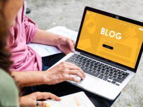 Two people sit outdoors, with one holding a laptop displaying a blog website with a yellow theme. The person's other hand is holding a pen and notebook. The background is blurred, focusing on the laptop screen that says "BLOG" in large white letters.