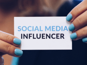 Close-up of a person holding a business card with neatly manicured hands. The card displays the text "SOCIAL MEDIA INFLUENCER" in capital letters, with "SOCIAL MEDIA" in blue and "INFLUENCER" in black. The person’s nails are painted a matching blue.