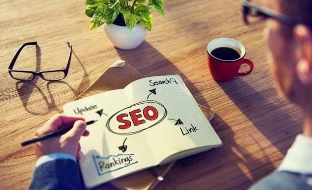 A person is sketching an SEO concept map on a notepad. The map includes terms like "Search," "Update," "Rankings," and "Link" connected to "SEO" in the center. On the table are eyeglasses, an open book, a potted plant, and a cup of coffee.