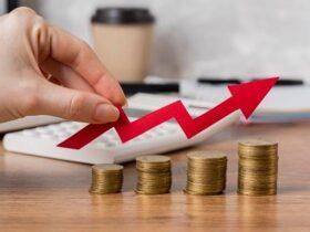 A hand holds a red upward-pointing arrow over four increasing stacks of coins, signifying financial growth. A calculator, coffee cup, and documents are blurred in the background on a wooden desk.
