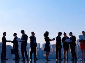 A diverse group of business professionals is standing in a line against a cityscape background. Some individuals are engaged in conversations, while others are examining documents or using electronic devices. The scene reflects a busy urban professional environment.