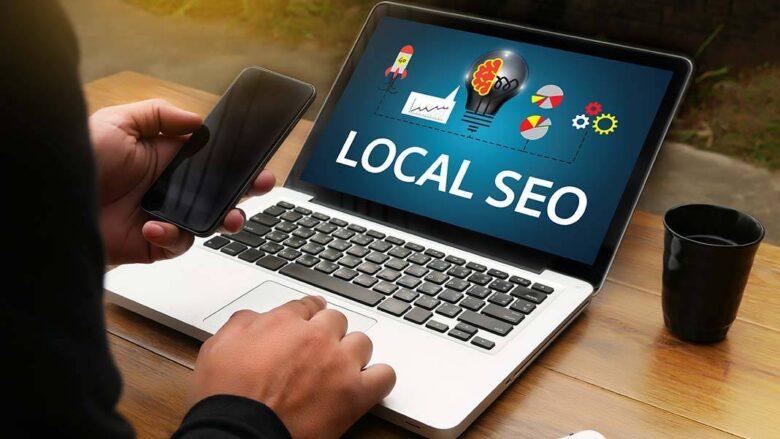 A person sits at a wooden table using a laptop displaying "LOCAL SEO" on the screen, with colorful icons representing digital marketing concepts. They hold a smartphone in their left hand and type on the laptop with their right. A black coffee cup is on the table.