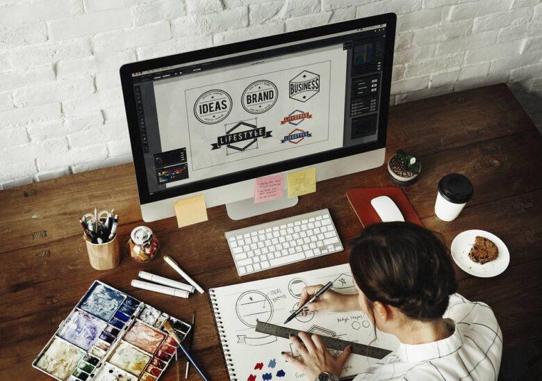 A person sits at a wooden desk, sketching in a notebook with art supplies scattered around. A large computer monitor displays various logo designs labeled "Ideas," "Brand," and "Business." The workspace includes a cup of coffee, watercolor paints, and a tablet.