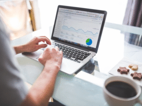 Person using a laptop displaying a graph and pie chart on the screen, indicative of data analysis or business analytics. There is a cup of coffee and a small plate with snacks next to the laptop on a glass-top table.