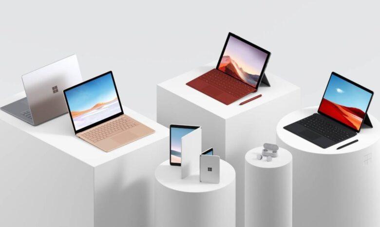 A collection of Microsoft Surface products displayed on white podiums against a gray background. The items include laptops, tablets with keyboards, earbuds, and a folding device. The products vary in colors including silver, bronze, and black.