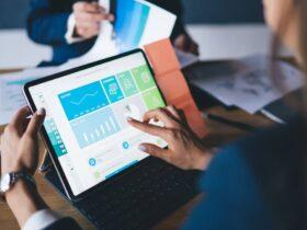 A person works on a tablet showing graphs and data charts, while another person in the background holds documents with graphs. Both are at a desk, indicating a business setting focused on data analysis or presentation.