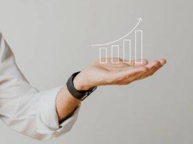 A person wearing a white shirt and a black smartwatch holds out their hand, which has a digitally overlaid, ascending bar graph with an arrow pointing upward, symbolizing growth and success. The background is plain and light-colored.