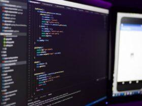 Two computer monitors are shown; the left screen displays code with syntax highlighting in an Integrated Development Environment (IDE), while the right screen shows a mobile application's interface. The workspace is dimly lit, highlighting the screens.