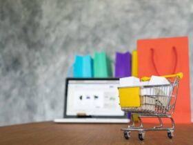 A miniature shopping cart filled with small boxes sits on a wooden surface. In the background, a laptop displaying a shopping website and several colorful shopping bags are visible. The scene depicts online shopping and e-commerce.
