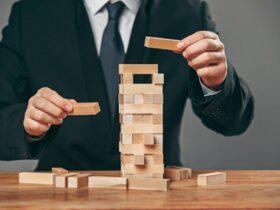 A person in a suit is building a tower from wooden blocks on a table. Their hands are placing a block on top of a partially constructed, unstable stack, with several blocks scattered around.