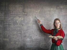 A person with long hair and glasses, wearing a red and green outfit, stands in front of a chalkboard covered in various diagrams and notes. They hold an open book in one hand and point to the chalkboard with a pointer in the other hand.