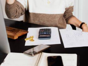 A person sitting at a desk with a brown and white sweater, partially visible. On the desk are a calculator, a wooden keyboard, keys, printed documents, and a smartphone. An open notebook and another smartphone are in the foreground.