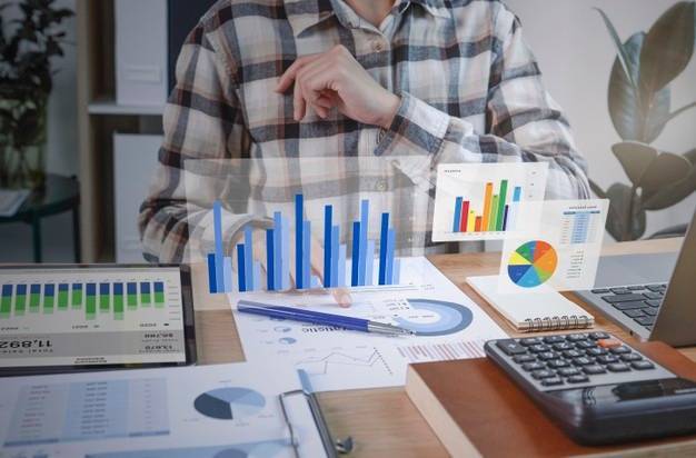 A person wearing a checkered shirt sits at a desk analyzing various charts and graphs on papers and electronic devices. A tablet, notebook, calculator, and other documents are on the desk. Holographic bar and pie charts are visibly displayed above the papers.