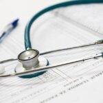 A stethoscope rests on top of a medical document with printed text, alongside a blue pen. The scene suggests a medical or healthcare setting, focusing on patient care or record-keeping.