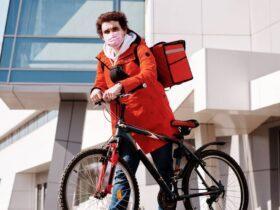A person wearing a red coat and a pink face mask is standing next to a black bicycle. They have a red delivery bag on their back and are in an urban area with modern buildings in the background.