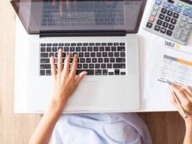 A person uses a laptop with their left hand, while the right hand holds a pen above a printed financial document on a desk. A calculator is visible to the right of the laptop, and the person appears to be working on financial data or accounting.