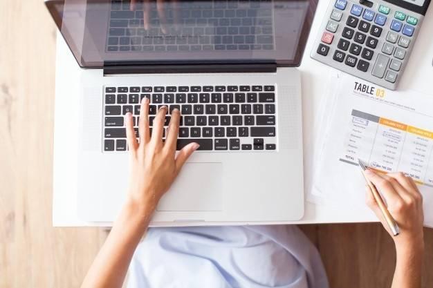 A person uses a laptop with their left hand, while the right hand holds a pen above a printed financial document on a desk. A calculator is visible to the right of the laptop, and the person appears to be working on financial data or accounting.