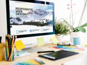 A well-organized workspace with a computer displaying a website about "Responsive Web Design." The desk has colorful pencils, sticky notes, glasses, a tablet, and color swatches. A potted plant adds a touch of greenery to the setup.