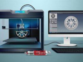 A 3D printer is producing a wheel design, as shown on a computer monitor next to it. A detailed rendering of the wheel is displayed on the monitor's screen. In front of the 3D printer and monitor, a red and silver mechanical component rests on the desk.