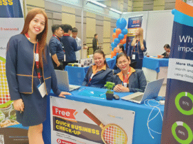 Three women at a digital marketing expo booth offering a free business check-up, with colorful banners, laptops, and a lively event background.