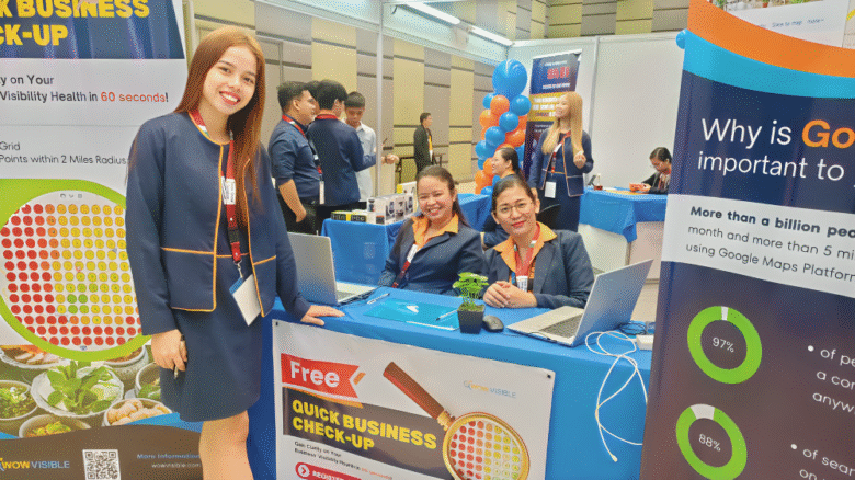 Three women at a digital marketing expo booth offering a free business check-up, with colorful banners, laptops, and a lively event background.