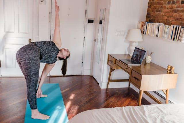 Woman practicing yoga at home on a blue mat while following an online class on her laptop placed on a wooden desk.