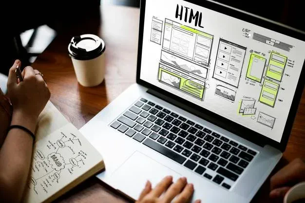 A person working on a laptop displaying HTML code and website wireframes on the screen. Another person is taking notes on a notepad with diagrams and notes visible. A cup of coffee is placed on the wooden desk beside them.