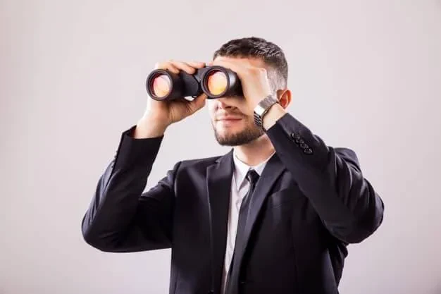 A man with short hair and a beard is wearing a dark suit and tie. He is holding a pair of binoculars up to his eyes and looking through them. The background is a plain, light gray color.