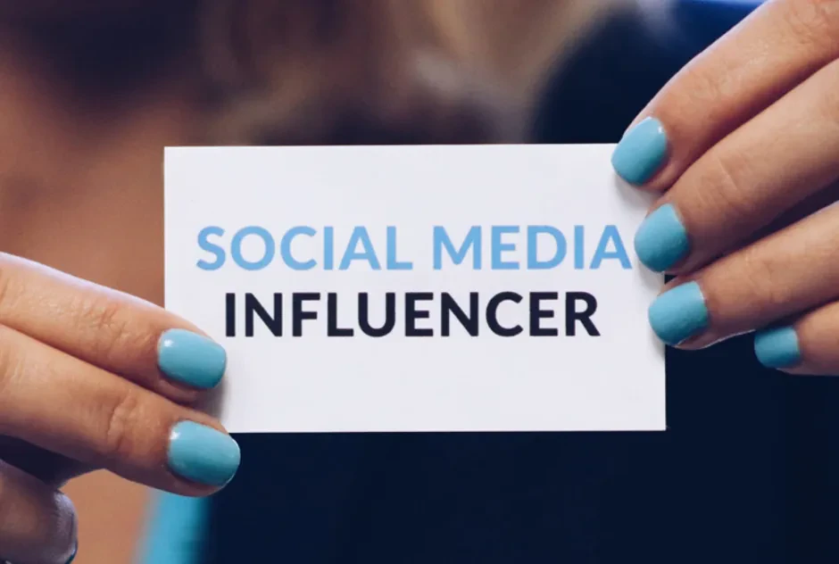 Close-up of a person holding a business card with neatly manicured hands. The card displays the text "SOCIAL MEDIA INFLUENCER" in capital letters, with "SOCIAL MEDIA" in blue and "INFLUENCER" in black. The person’s nails are painted a matching blue.