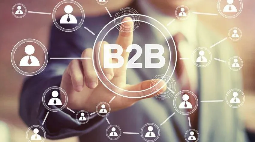 A person in a suit is pressing a virtual button labeled "B2B" on a transparent screen. The screen is filled with interconnected icons of people, symbolizing a business-to-business network. The background is blurred to highlight the interaction.