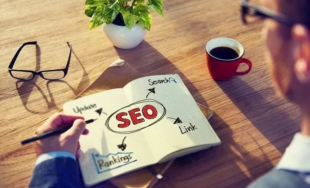 A person is sketching an SEO concept map on a notepad. The map includes terms like "Search," "Update," "Rankings," and "Link" connected to "SEO" in the center. On the table are eyeglasses, an open book, a potted plant, and a cup of coffee.