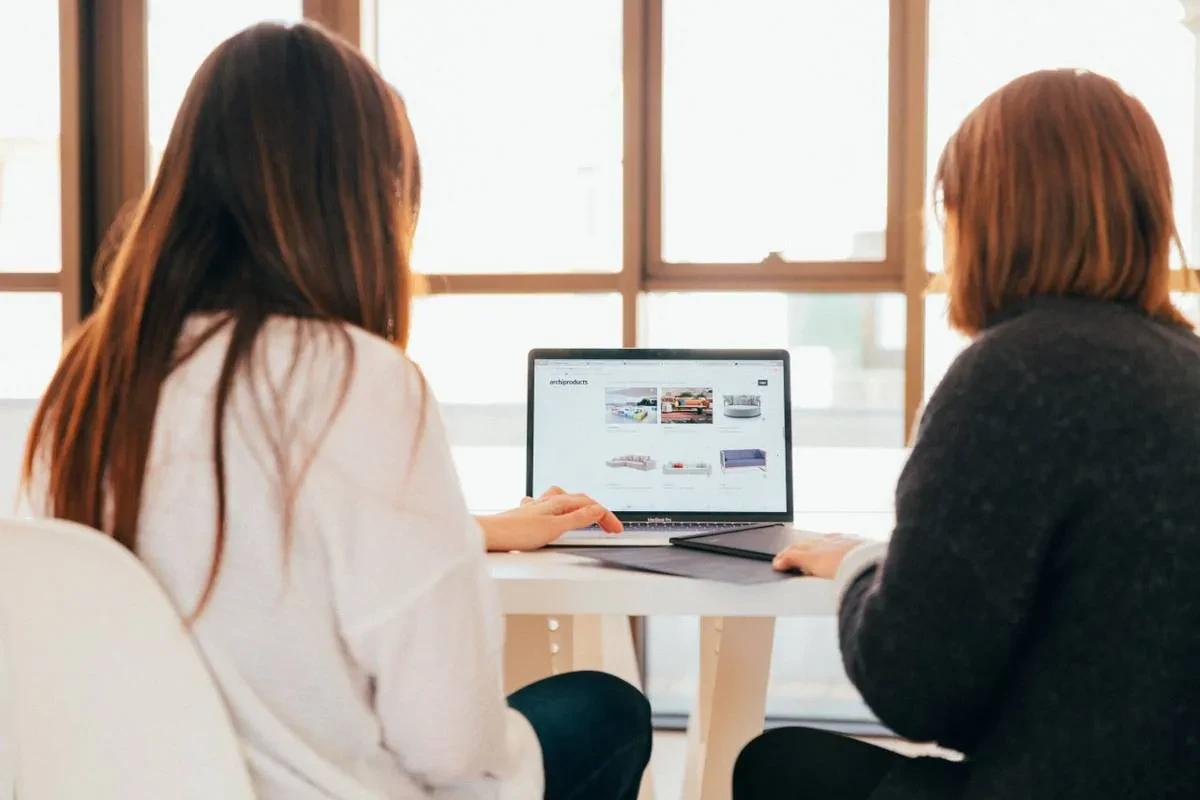 Two people are sitting at a table, facing a laptop. The screen shows a travel website with various travel-related images and options. They appear to be discussing or planning something, possibly a trip. The room is well-lit by large windows in the background.