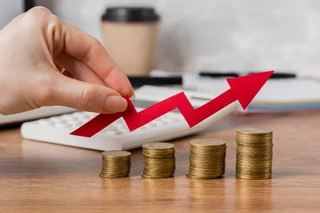 A hand holds a red upward-pointing arrow over four increasing stacks of coins, signifying financial growth. A calculator, coffee cup, and documents are blurred in the background on a wooden desk.