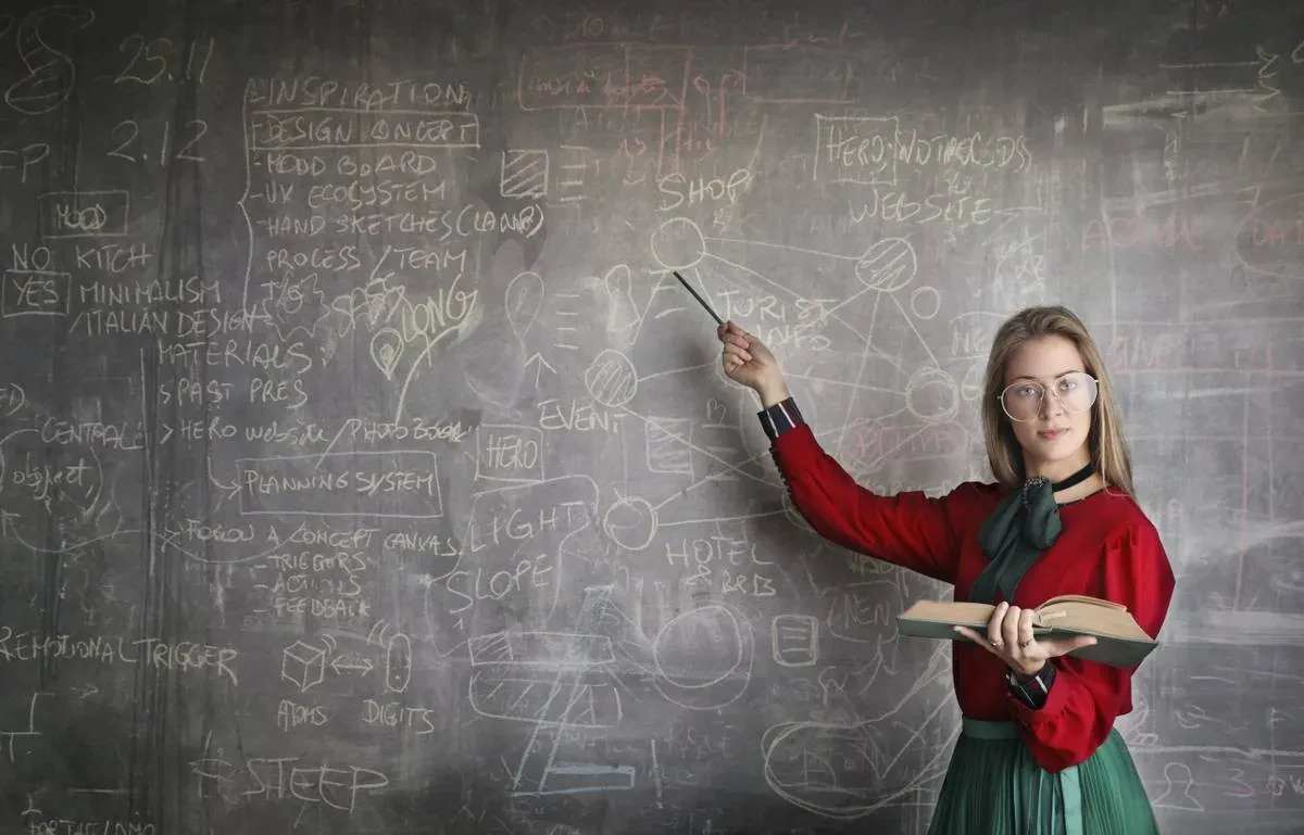 A person with long hair and glasses, wearing a red and green outfit, stands in front of a chalkboard covered in various diagrams and notes. They hold an open book in one hand and point to the chalkboard with a pointer in the other hand.