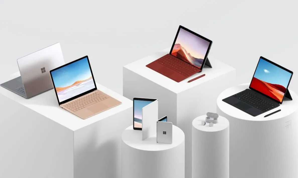 A collection of Microsoft Surface products displayed on white podiums against a gray background. The items include laptops, tablets with keyboards, earbuds, and a folding device. The products vary in colors including silver, bronze, and black.