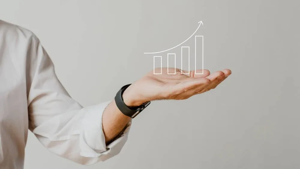 A person wearing a white shirt and a black smartwatch holds out their hand, which has a digitally overlaid, ascending bar graph with an arrow pointing upward, symbolizing growth and success. The background is plain and light-colored.